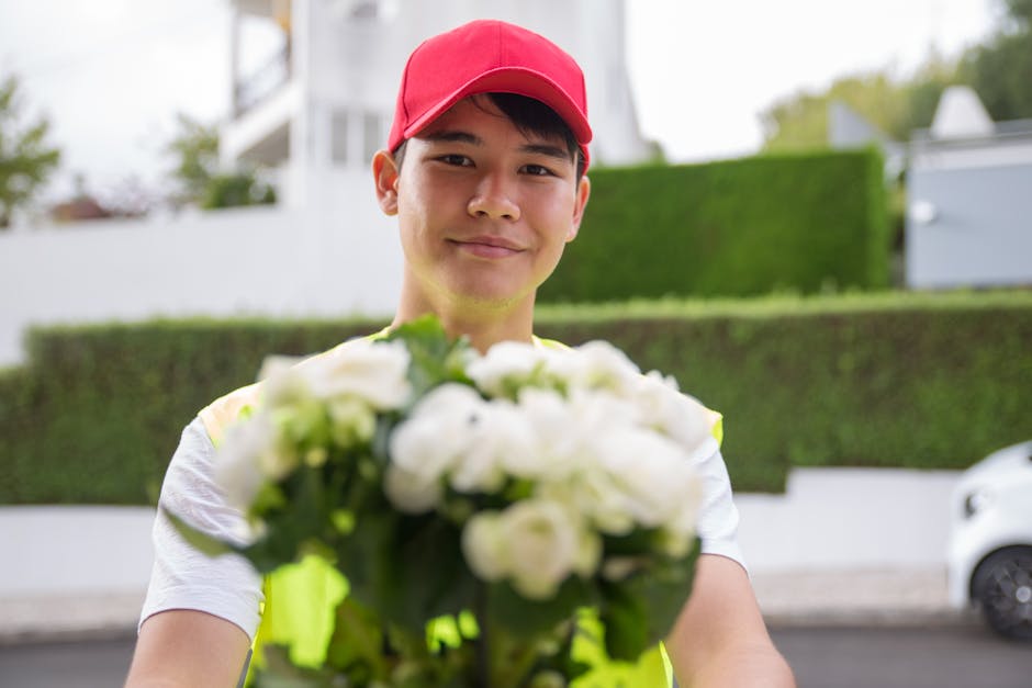 A delivery person wearing a red cap and a high-visibility vest is holding a bouquet of fresh white roses and green foliage. The bouquet is wrapped in clear plastic and is positioned toward the camera,
