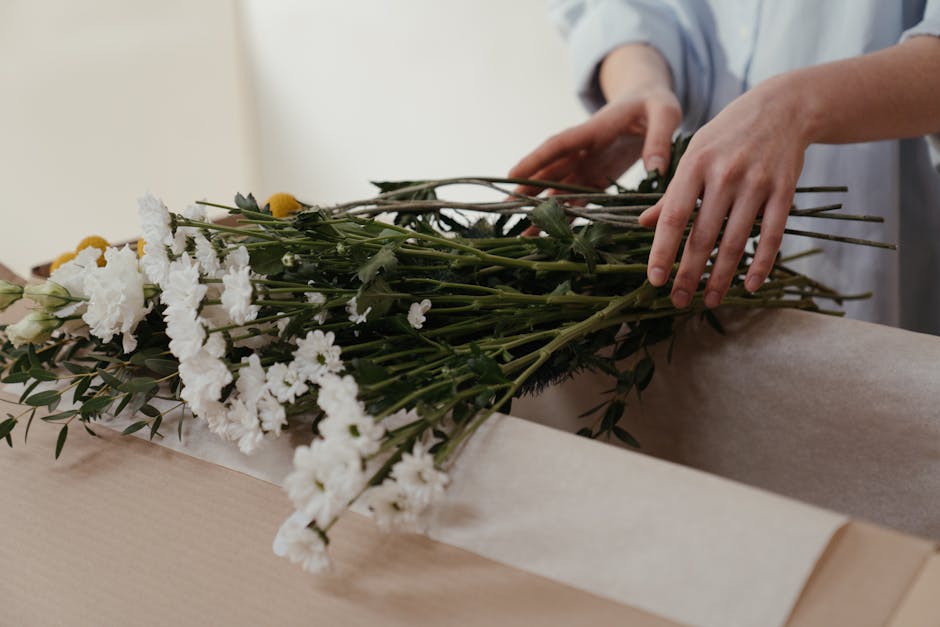 A person carefully arranging a bouquet of fresh white carnations, yellow billy balls, and green foliage on a light-colored wooden table, preparing it for flower delivery from Hatton Florist. The bouqu