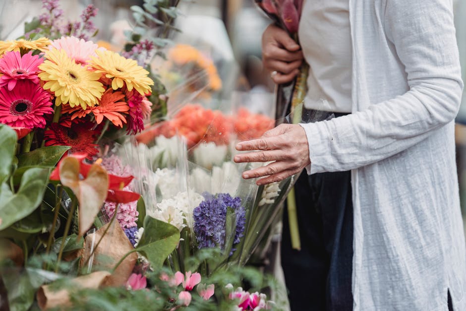 A person dressed in a white long-sleeve top and dark pants browsing a flower stall on a street market, selecting floral arrangements. The display features a variety of fresh flowers, including yellow,