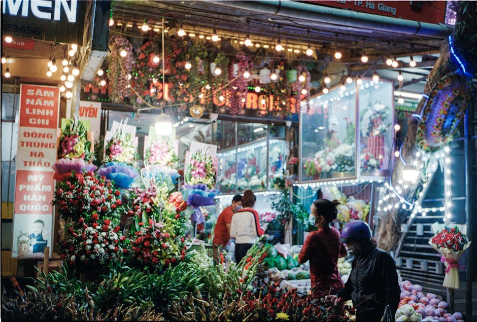 A vibrant flower market scene illuminated by colorful string lights and decorative lanterns, featuring numerous floral bouquets and arrangements in vivid reds, pinks, purples, and whites. The display