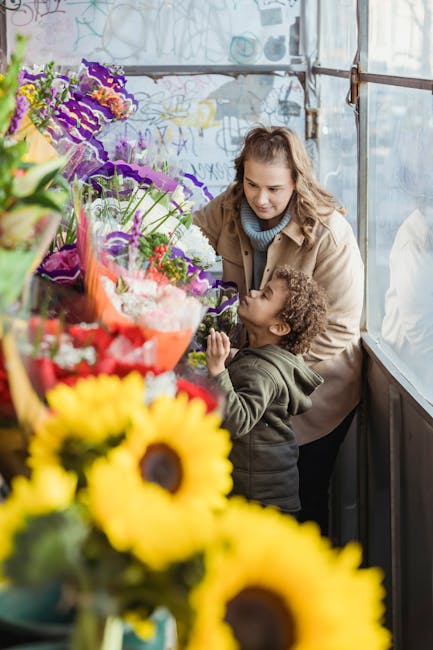 A woman and a young girl are inside a flower shop, inspecting a vibrant bouquet of seasonal flowers. The arrangement includes bright red roses, purple irises, and other colorful blooms wrapped in clea