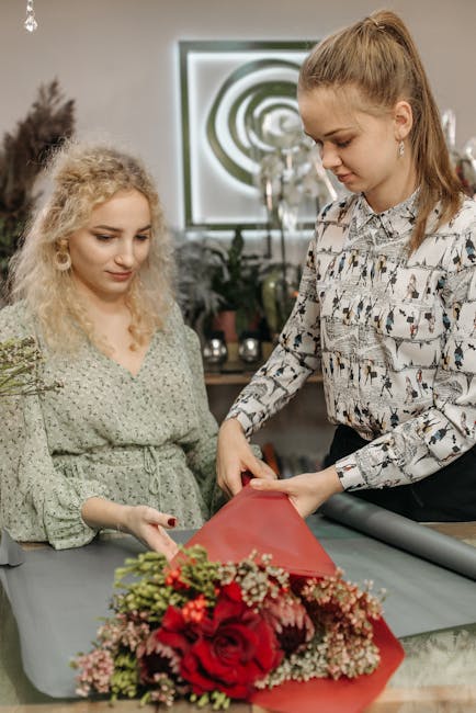 A professional florist from Hatton Florist is wrapping a vibrant bouquet of red roses, pink and white baby's breath, and assorted greenery in red craft paper. The arrangement is being prepared on a ta
